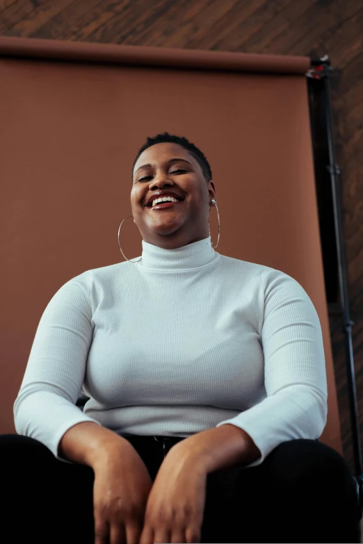 a woman sitting in a chair with a smile on her face, an album cover, trending on unsplash, full figured, in white turtleneck shirt, non binary model, thicc build