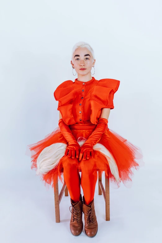 a woman in a red dress sitting on a chair, an album cover, featured on reddit, antipodeans, fluffy orange skin, non binary model, avant designer uniform, with a white complexion