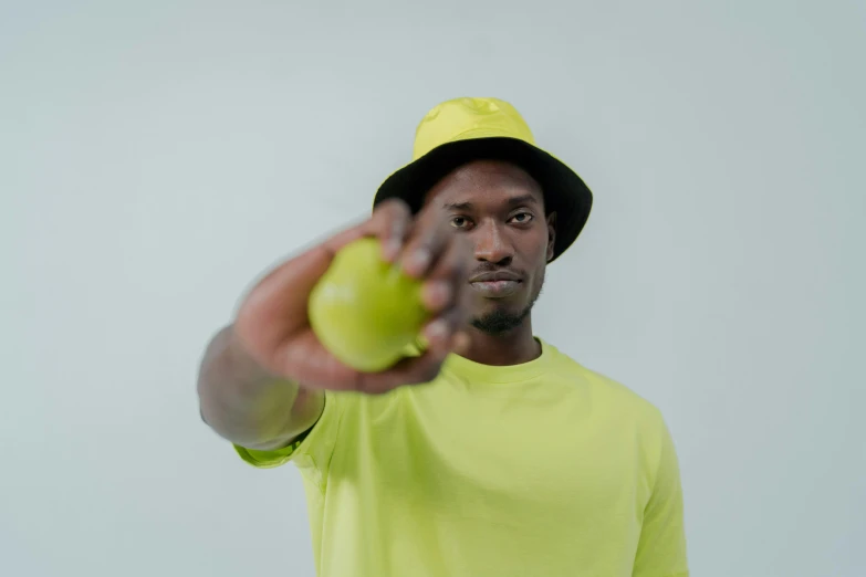 a man in a yellow shirt holding a green apple, by Barthélemy Menn, unsplash, lyco art, wearing a baseball cap, pointing at the camera, adut akech, on a pale background