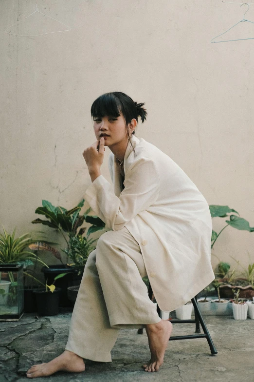 a woman sitting on a chair talking on a cell phone, an album cover, inspired by Ruth Jên, unsplash, wearing white pajamas, indonesia, portrait androgynous girl, plants