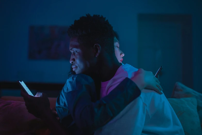 a man and a woman sitting on a bed looking at a cell phone, pexels, pink and blue lighting, two men hugging, dark skin, netflix