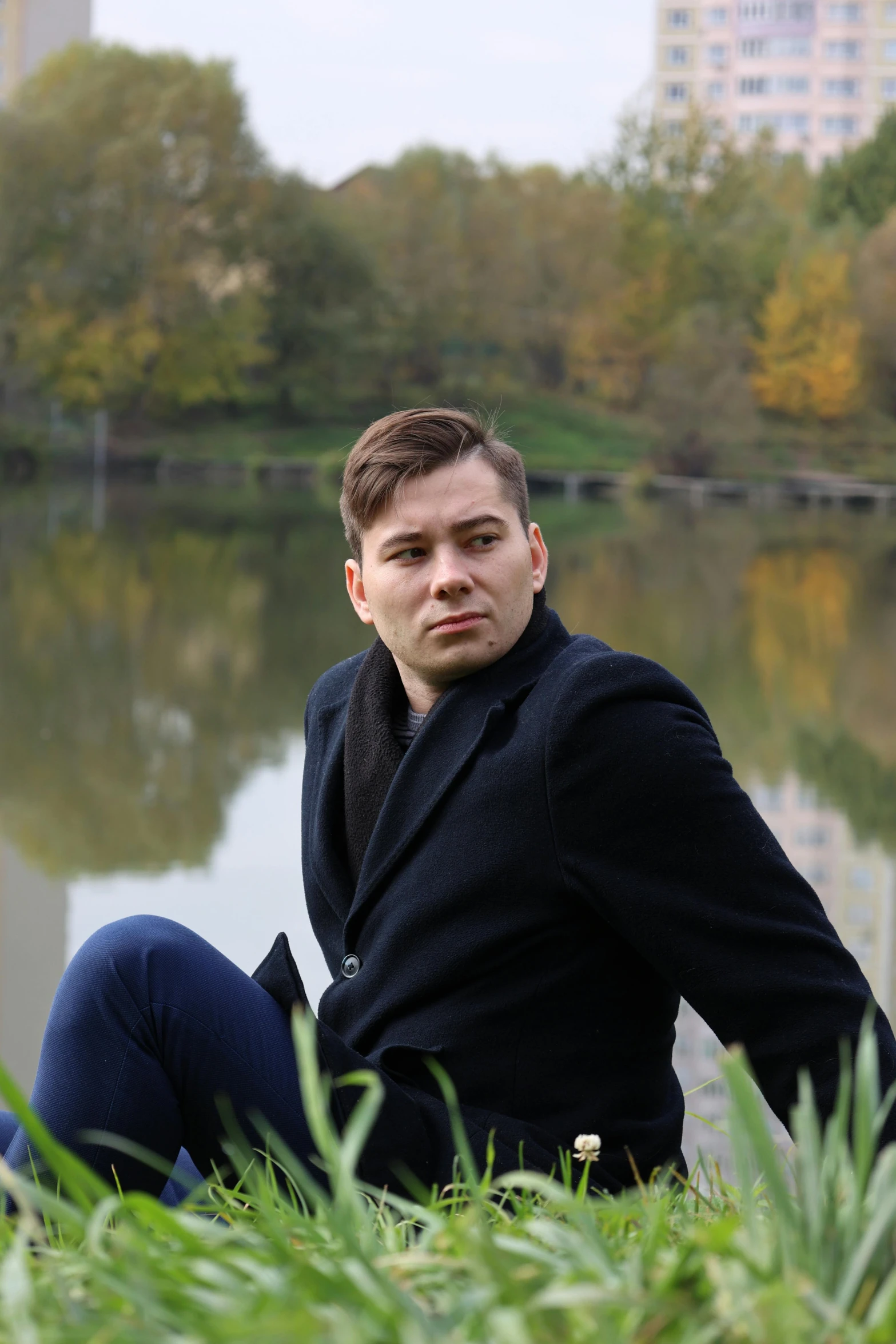 a man sitting in the grass next to a body of water, an album cover, by Alexander Fedosav, reddit, wearing a fancy black jacket, andrea savchenko, 15081959 21121991 01012000 4k, headshot profile picture