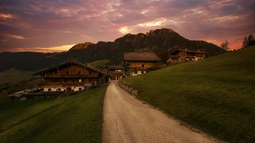 a dirt road running through a lush green hillside, by Sebastian Spreng, pexels contest winner, renaissance, wooden houses, sundown, austria, epic mountains in the background