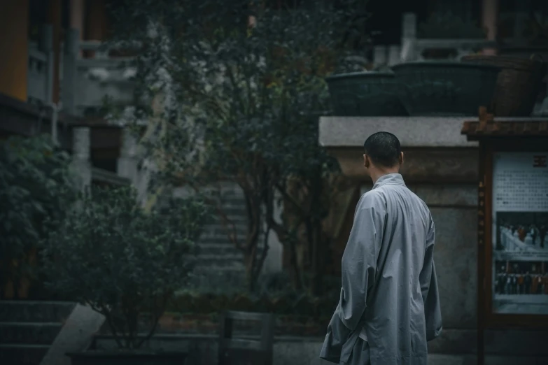 a man in a robe walking down a street, inspired by Ma Quan, pexels contest winner, grey clothes, contemplating, standing over a tomb stone, bo xun ling