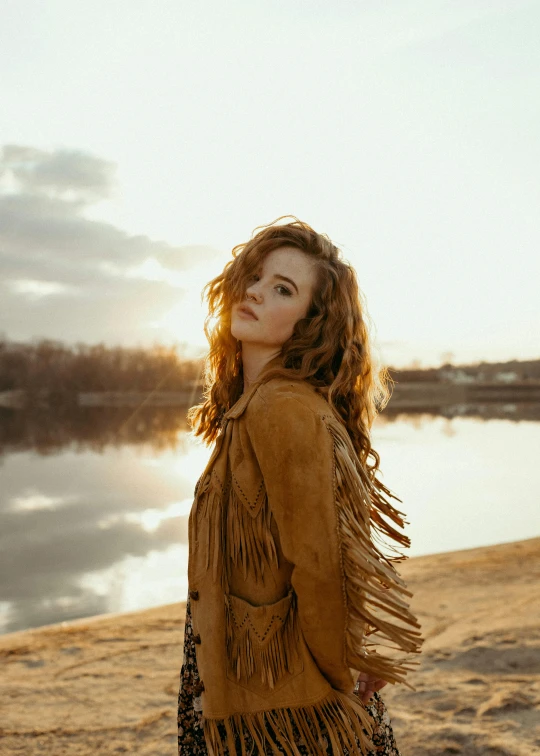 a woman standing on a beach next to a body of water, an album cover, pexels contest winner, photorealism, ginger wavy hair, wearing brown leather jacket, promotional image, soft backlighting