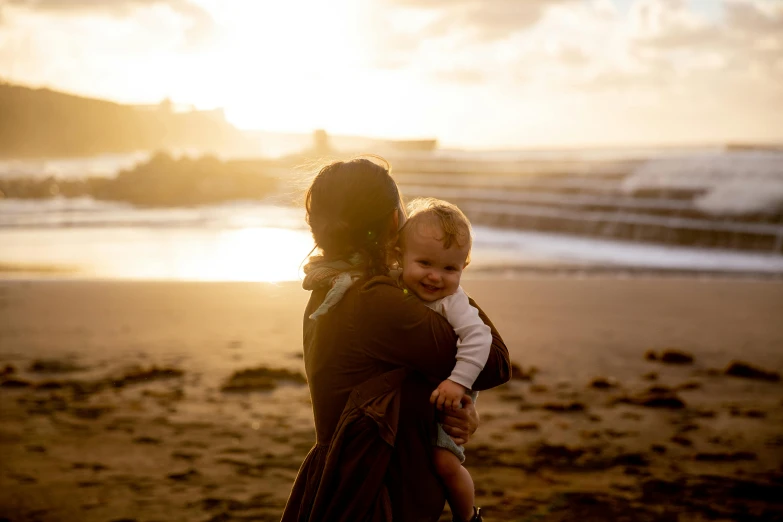 a woman holding a baby on a beach, by Matt Stewart, pexels contest winner, evening sunlight, looking happy, brown, instagram post