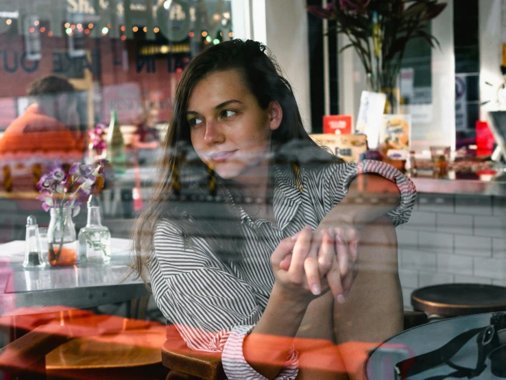 a woman sitting at a table in front of a window, by Julia Pishtar, pexels contest winner, inside in a glass box, sydney hanson, inside a french cafe, very reflective