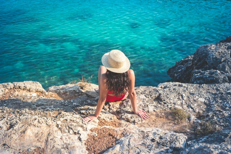 a woman sitting on top of a rock next to the ocean, red hat, turquoise water, avatar image