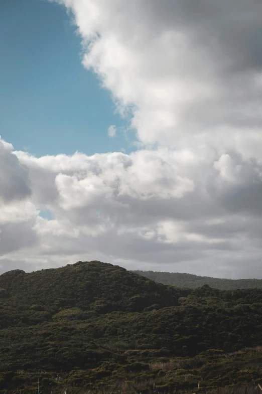 a man flying a kite on top of a lush green hillside, by Jacob Toorenvliet, unsplash, australian tonalism, layered stratocumulus clouds, seen from a distance, light blue sky with clouds, rocky hills