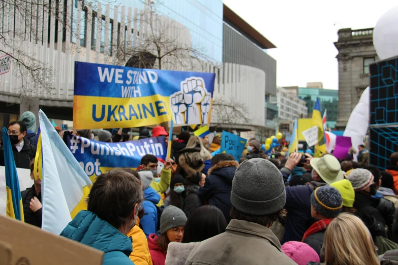 a group of people that are standing in the street, ukrainian flag on the left side, yellow and blue and cyan, protesters holding placards, canada