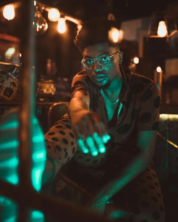 a man that is sitting down in a room, trending on pexels, afrofuturism, dimly lit dive bar, bisexual lighting, man with glasses, holding it out to the camera