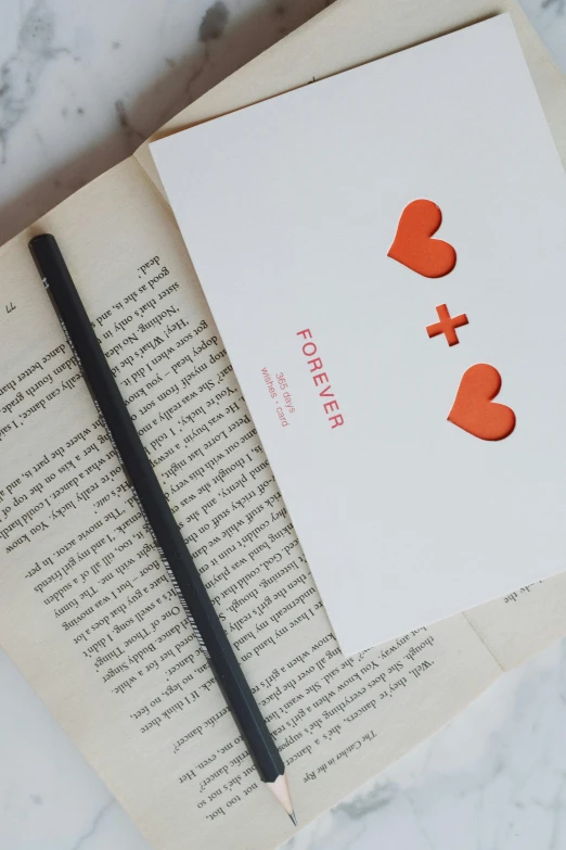 an open book sitting on top of a table next to a pencil, an album cover, by Julia Pishtar, pexels contest winner, private press, hearts symbol, orange and white color scheme, two people, focus on card