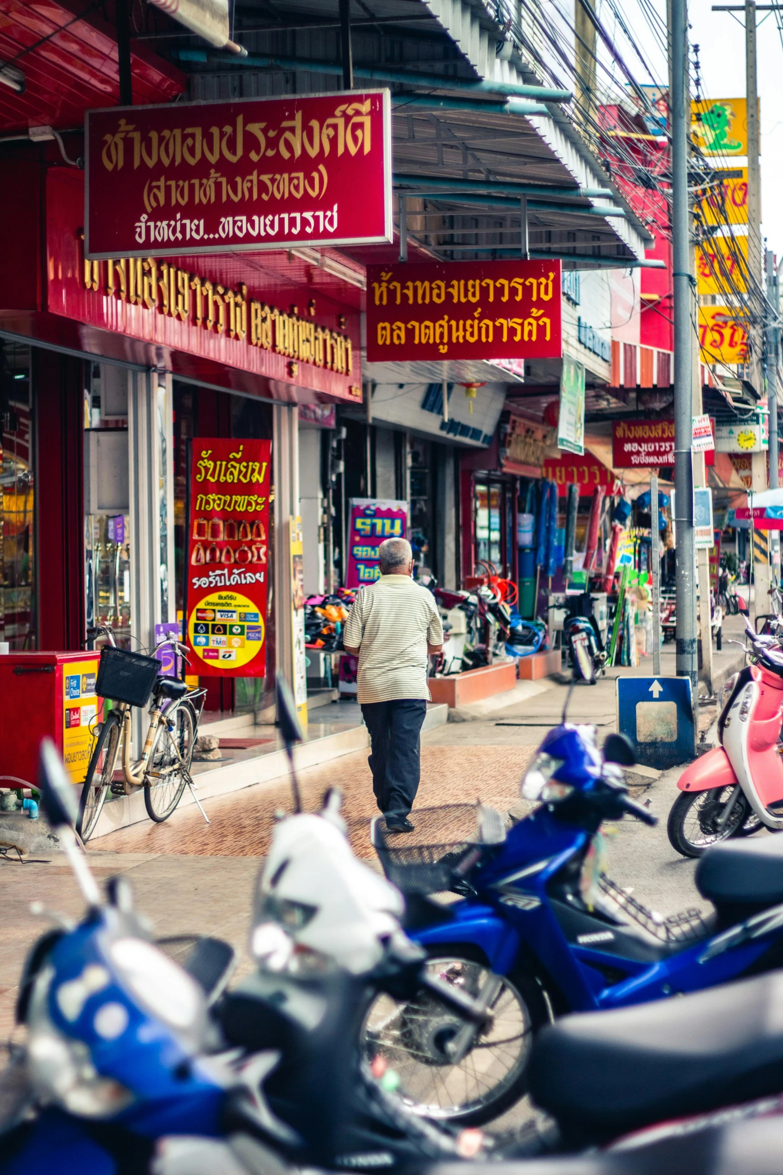 a group of motorcycles parked on the side of a street, patiphan sottiwilai, lots of signs and shops, man walking through city, vibrant colour