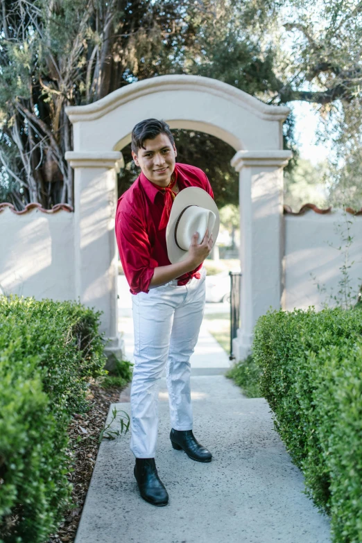 a man in a red shirt and white pants holding a hat, an album cover, inspired by Byron Galvez, in garden, androgynous male, david marquez, doing an elegant pose