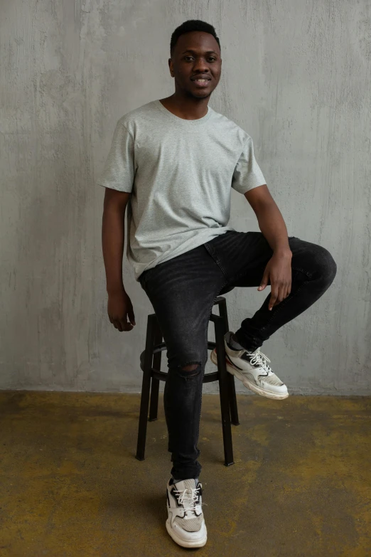 a man sitting on a stool in front of a wall, trending on pexels, jeans and t shirt, in gunmetal grey, riyahd cassiem, full - body - front - shot