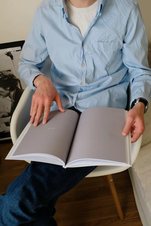 a man sitting in a chair reading a book, by David Simpson, photorealism, book in hand, wearing a light shirt, grey, colour photograph