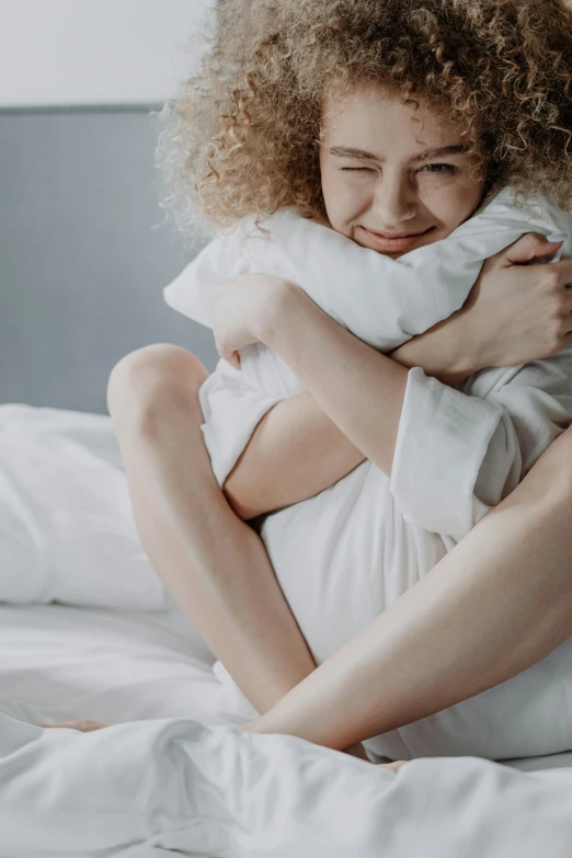 a woman sitting on top of a bed holding a pillow, trending on pexels, hugging each other, curly haired, gif, feels good man