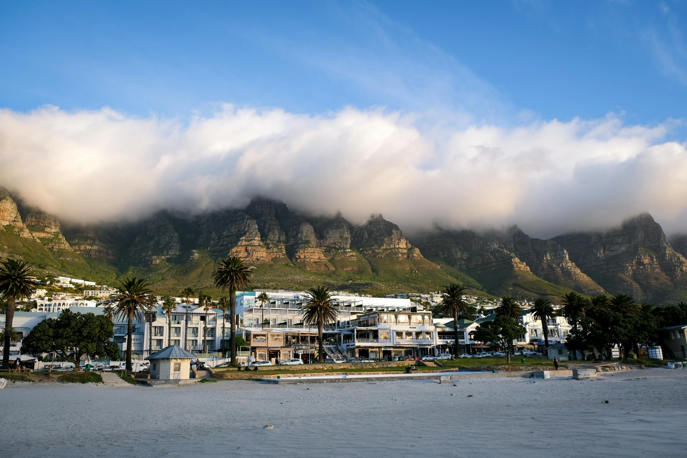 a view of a beach with mountains in the background, by Tom Wänerstrand, pexels contest winner, whitewashed buildings, royal cape, in a cloud, late afternoon