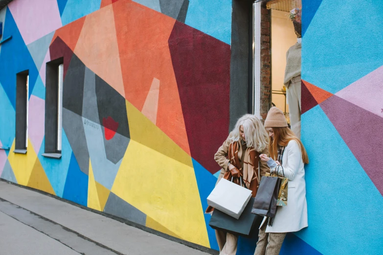 a couple of women standing next to a colorful building, by Julia Pishtar, pexels contest winner, street art, people shopping, ad image, cardboard