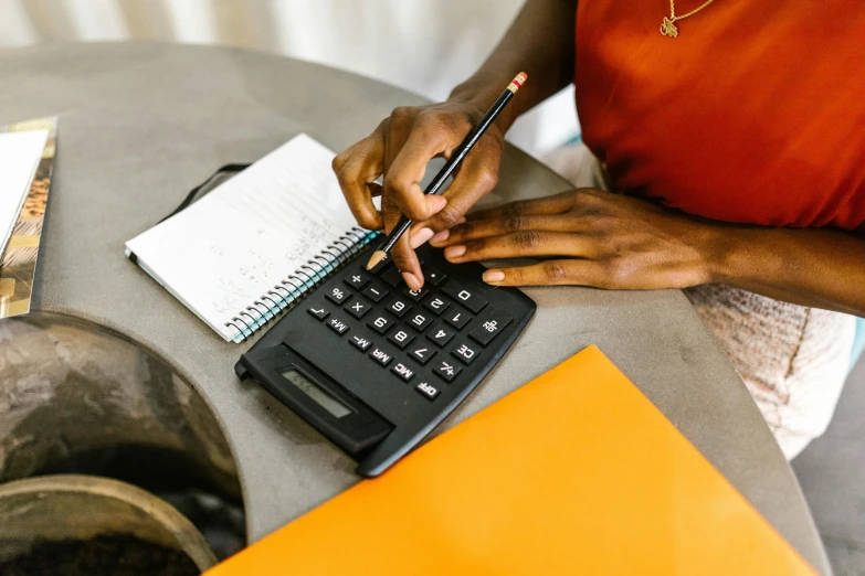 a woman sitting at a table using a calculator, by Meredith Dillman, trending on pexels, photo of a black woman, black and orange colour palette, educational supplies, holding notebook