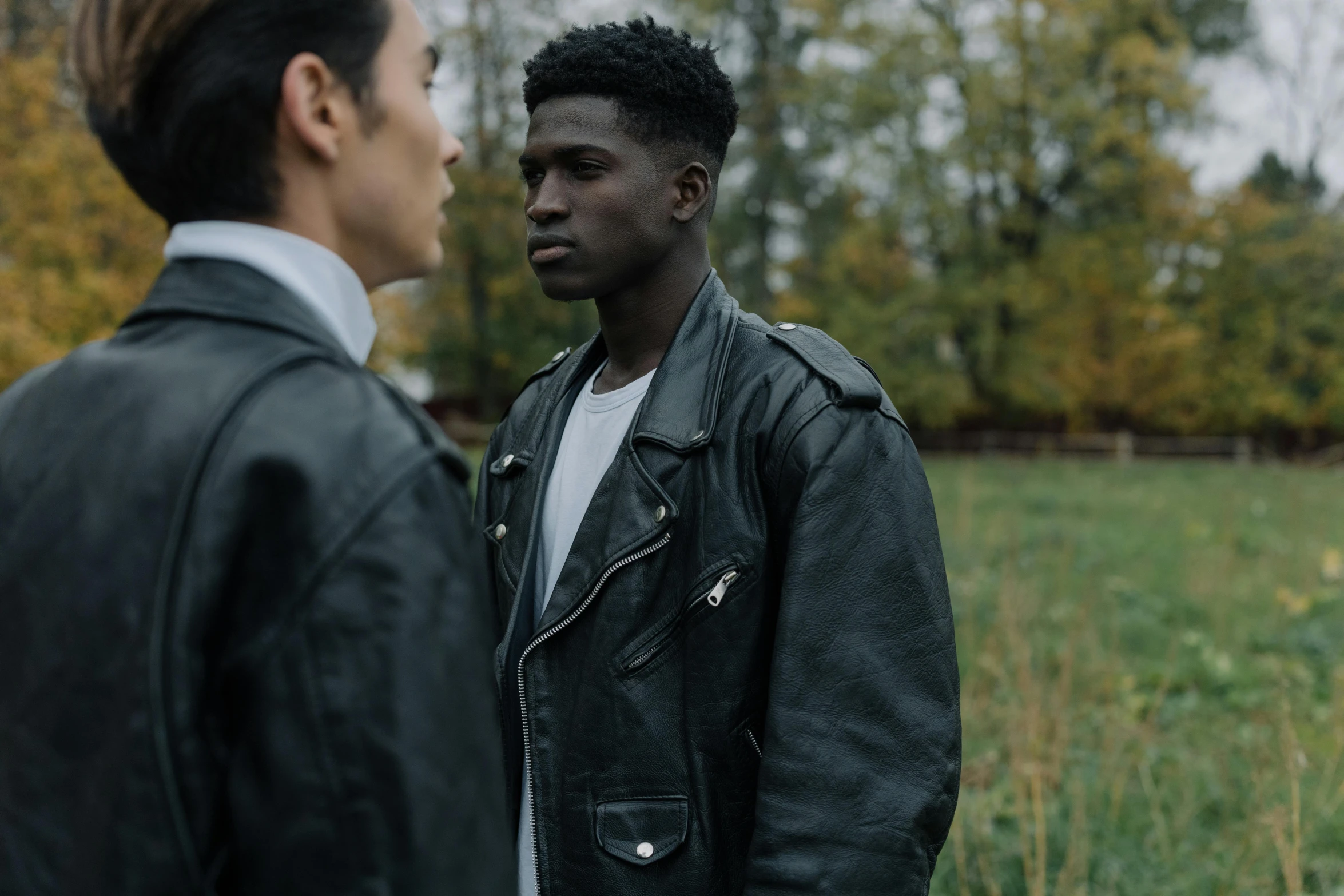 two men standing next to each other in a field, trending on pexels, visual art, black leather jacket, black teenage boy, looking at each other mindlessly, androgynous person