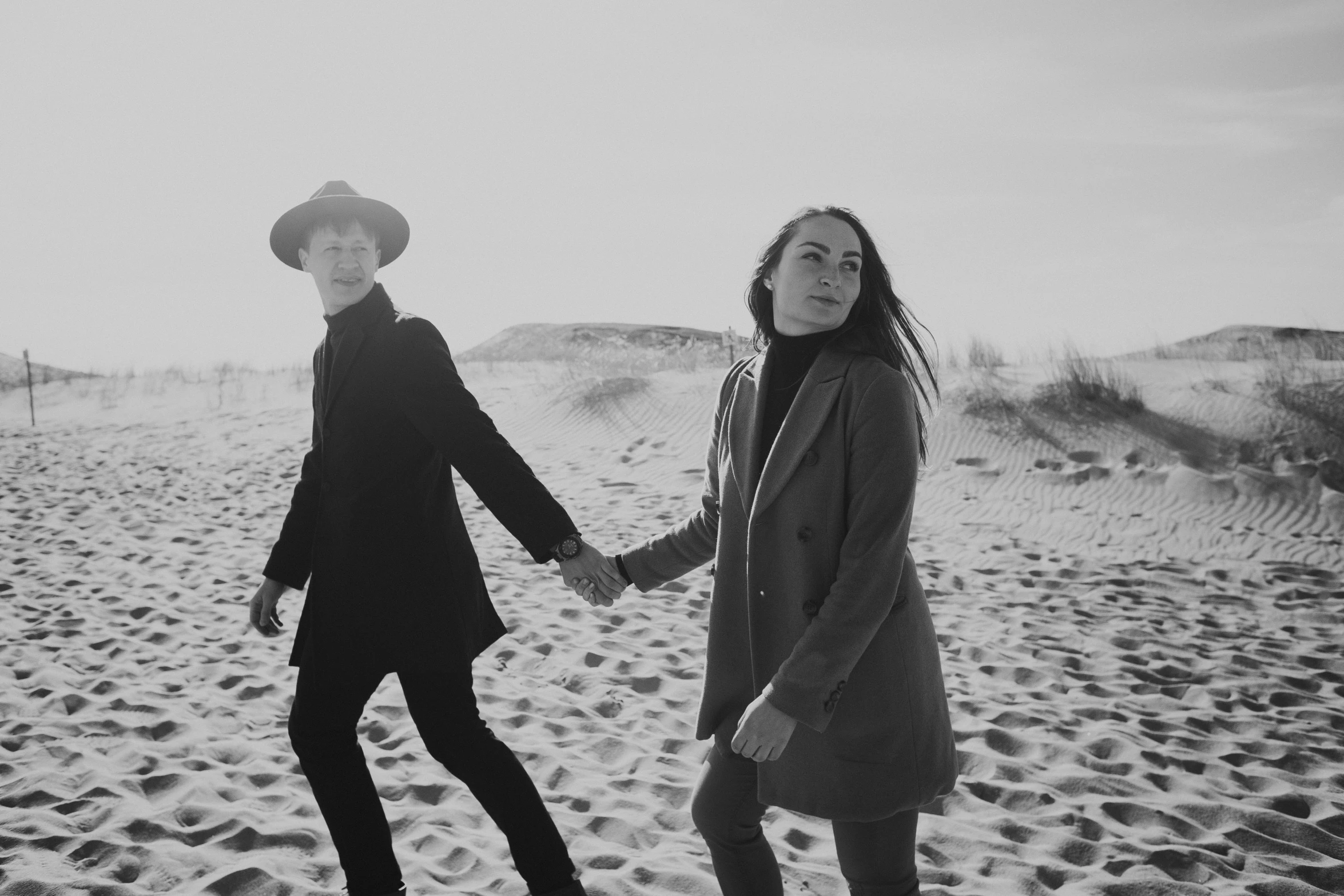 a black and white photo of two people holding hands, by Emma Andijewska, walking over sand dunes, straw hat and overcoat, ska skeleton and girlfriend, chillwave