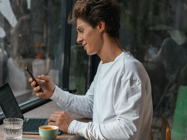 a man sitting at a table with a laptop and a cell phone, trending on pexels, happening, avatar image, cute young man, lachlan bailey, high quality picture