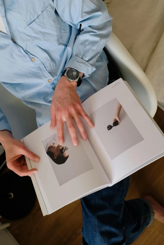 a man sitting in a chair holding a book, an album cover, by Nina Hamnett, unsplash, photorealism, on a white table, hands shielding face, watch photo, printed page