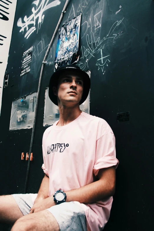 a man sitting in front of a wall with graffiti on it, an album cover, unsplash contest winner, pink shirt, wearing a chocker and cute hat, luka mivsek, standing with a black background