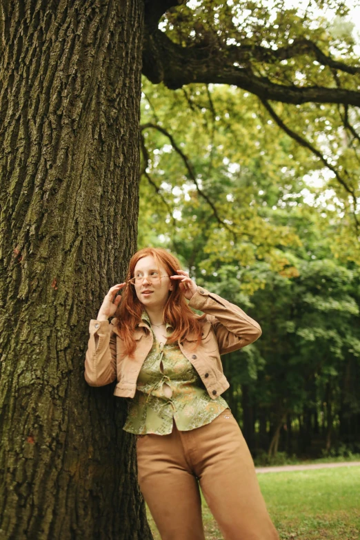 a woman leaning against a tree in a park, an album cover, inspired by Elsa Bleda, trending on pexels, renaissance, hr ginger, green corduroy pants, wearing jacket, young woman looking up