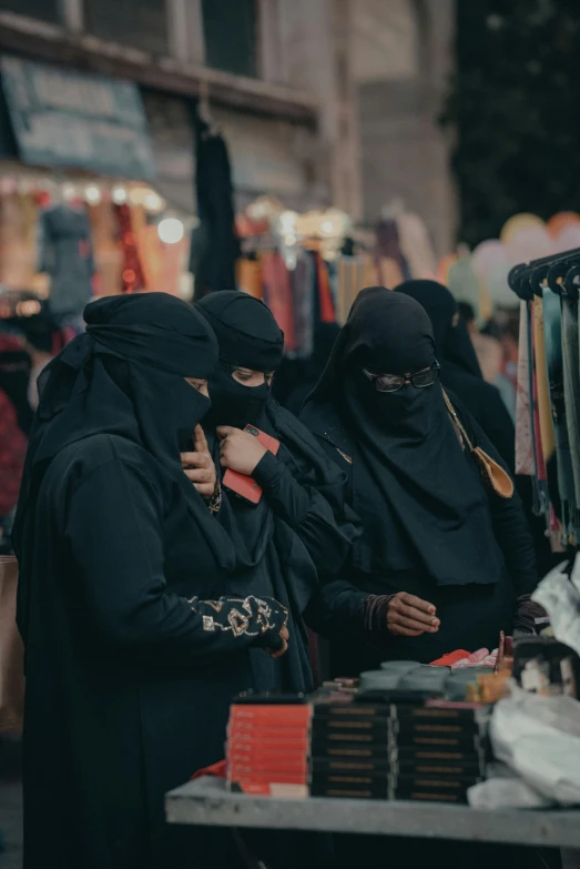 a group of women standing next to each other, by Ibrahim Kodra, trending on unsplash, hurufiyya, dressed in a black cloak, marketplace, he wears dark visors, arabia