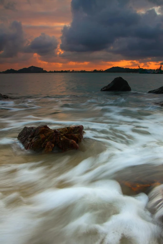 a sunset over a body of water with rocks in the foreground, by Dan Scott, romanticism, turbulent waves, vietnam, fujifilm”