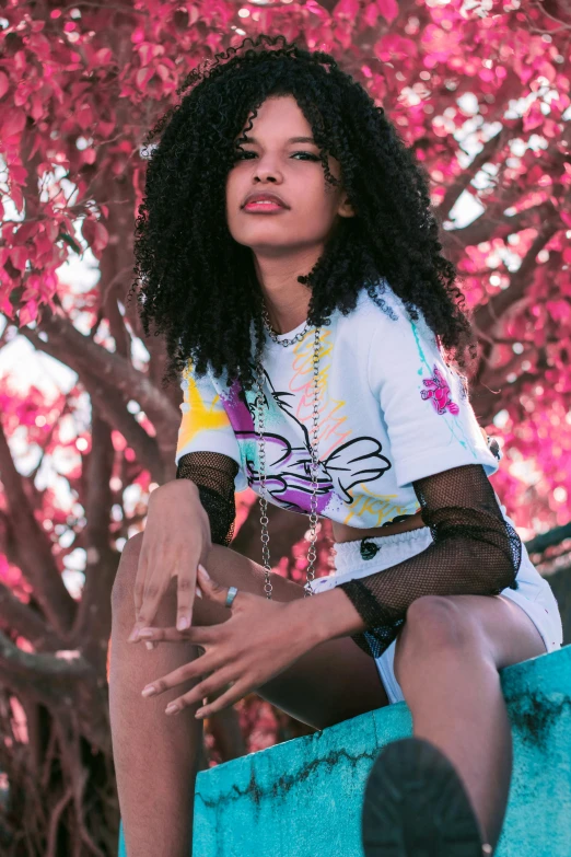 a woman sitting on top of a wall next to a tree, trending on pexels, graffiti, long afro hair, graphic tees, pastel goth aesthetic, woman in flowers