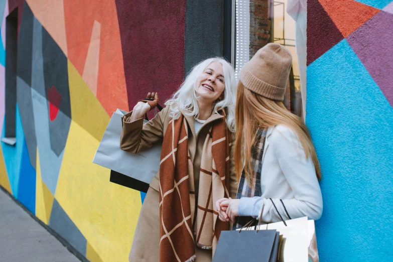 two women standing next to a colorful wall with shopping bags, trending on pexels, fine art, older woman, a blond, wearing a brown cape, smiling playfully