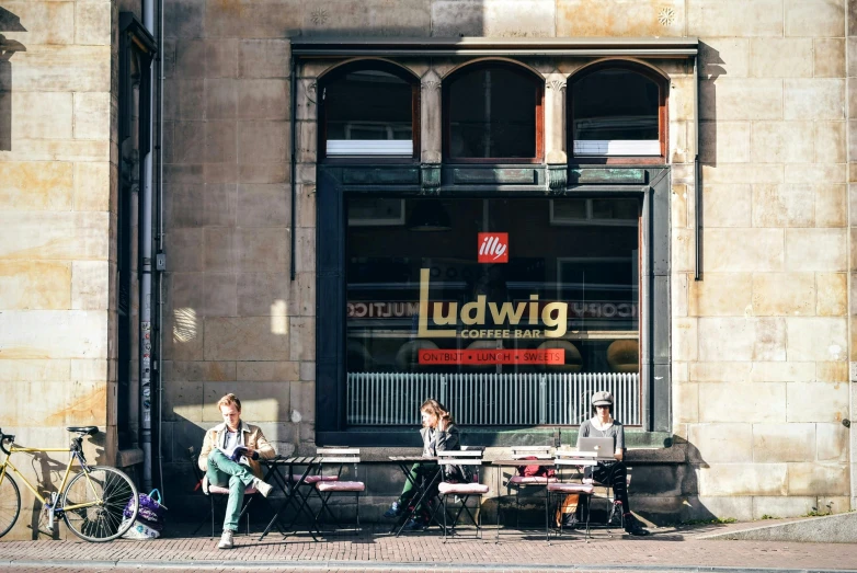 a group of people sitting at a table in front of a building, by Lodewijk Bruckman, trending on unsplash, award winning shopfront design, ludwig hohlwein, demur, in a square