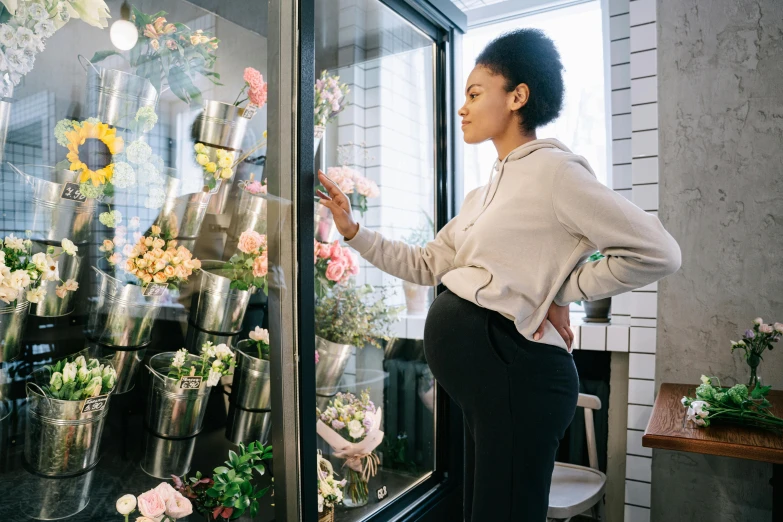 a woman standing in front of a display of flowers, trending on unsplash, pregnant belly, looking in the window, customers, inspect in inventory image