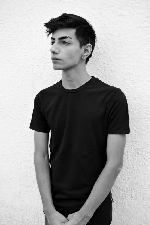 a man standing in front of a white wall, a black and white photo, inspired by Jean Malouel, pexels contest winner, antipodeans, wearing a black tshirt, 1 7 - year - old boy thin face, skinny caucasian young man, shabab alizadeh