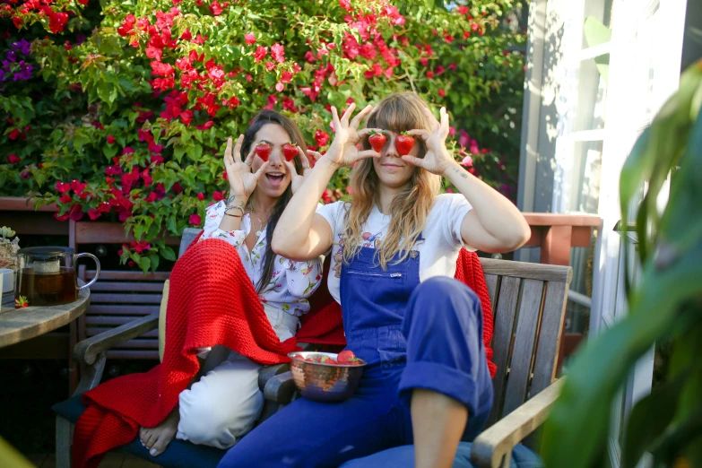 a couple of women sitting on top of a wooden bench, by Julia Pishtar, pexels, red sunglasses, with fruit trees, wearing overalls, red and blue garments