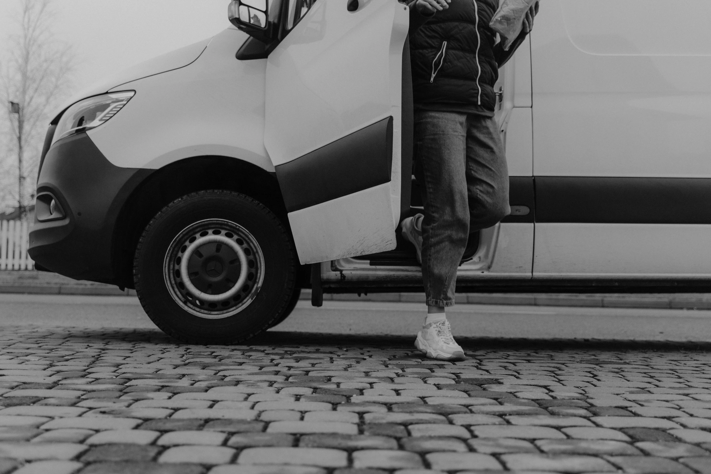 a black and white photo of a man standing next to a van, pexels contest winner, wearing white sneakers, background image, moving, high quality upload