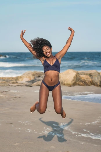 a woman jumping in the air on a beach, by Carey Morris, matte navy - blue bodysuit, wearing two - piece swimsuit, dark skin tone, smiling confidently