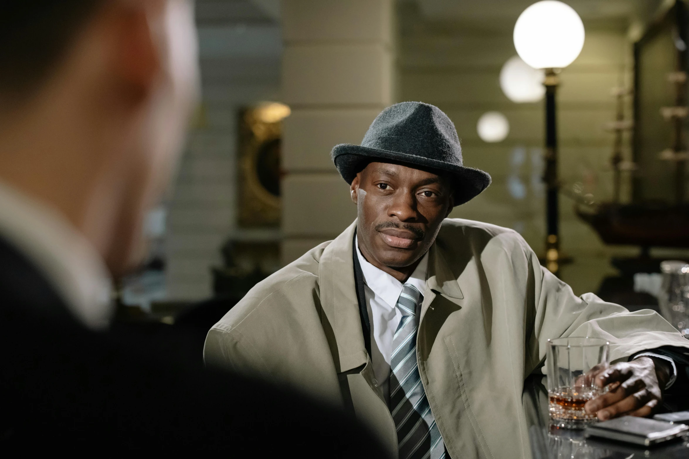 a man sitting at a bar with a drink in his hand, inspired by Charles Alston, lance reddick, a suited man in a hat, colour portrait photograph, ( ( theatrical ) )