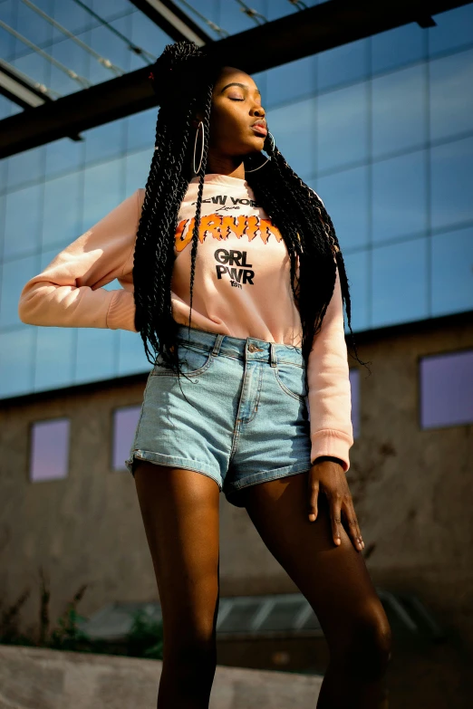 a woman standing on a skateboard in front of a building, by Quinton Hoover, trending on pexels, long black braids, toned orange and pastel pink, denim shorts, wearing sweatshirt