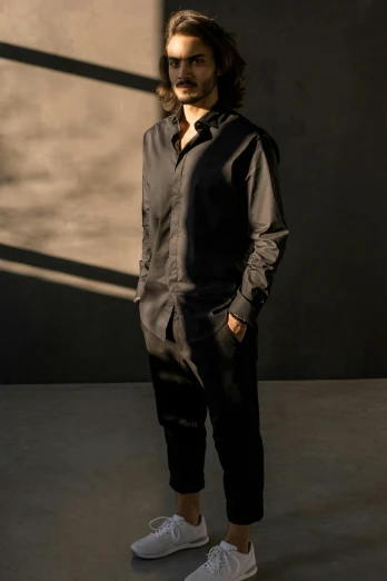 a man with long hair standing in front of a wall, jumpsuit, wearing a black shirt, style of maciej kuciara, f / 2 0