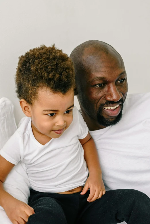 a man sitting next to a little boy on a bed, an album cover, by Samuel Washington Weis, pexels contest winner, balding hair, youthful taliyah, african, on a couch