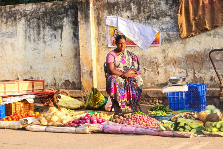 a woman sitting in front of a pile of fruit and vegetables, by Ingrida Kadaka, pexels contest winner, on an indian street, square, brown, thumbnail