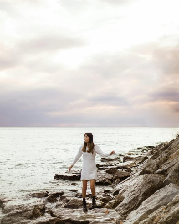 a woman standing on a rocky beach next to the ocean, by Julia Pishtar, unsplash, wearing a white hospital gown, lgbtq, teenage girl, multiple stories