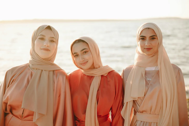 three women standing next to each other on a beach, inspired by Maryam Hashemi, trending on pexels, hurufiyya, flowing salmon-colored silk, white hijab, tan complexion, women's faces