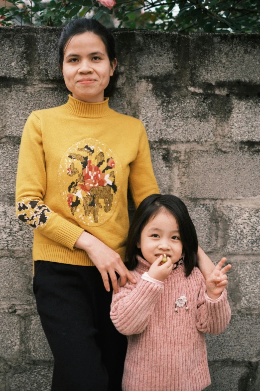 a woman and a little girl standing next to a stone wall, inspired by Zhang Kechun, pexels contest winner, pixel art, wearing yellow floral blouse, wearing a sweater, film portrait, promotional image