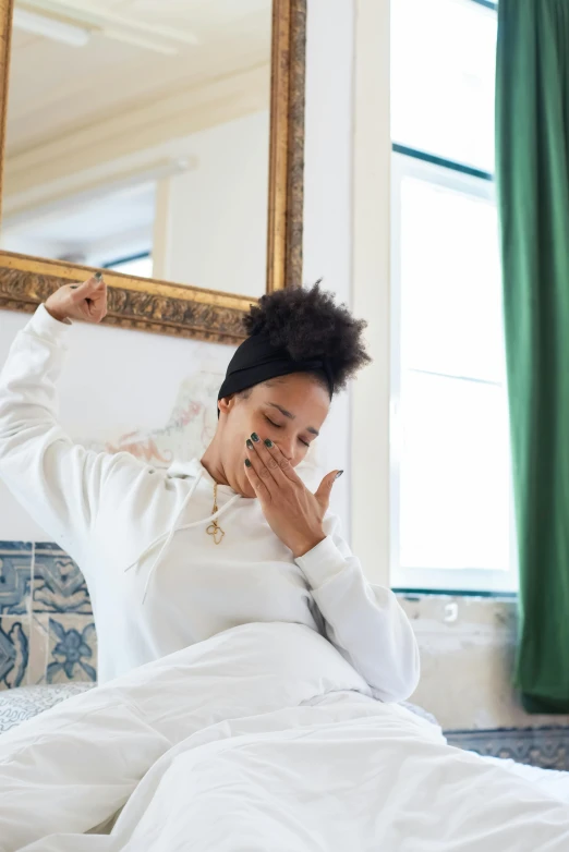 a woman sitting on top of a bed covered in white sheets, by Dulah Marie Evans, trending on pexels, happening, yawning, nathalie emmanuel, wearing a white sweater, gold hour