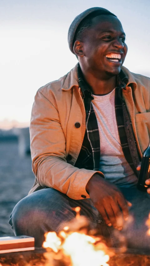 a man sitting next to a campfire holding a bottle of wine, trending on pexels, renaissance, brown jacket, african american, sunfaded, happy friend